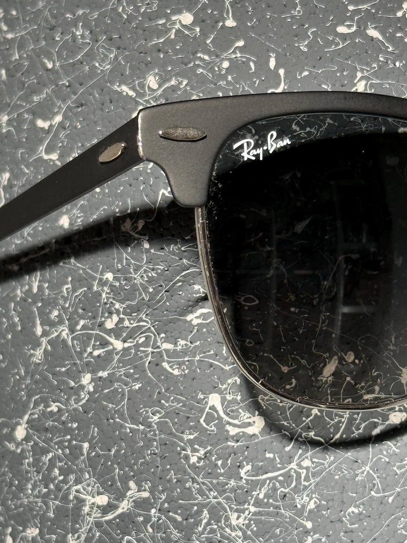 May include: Black Ray-Ban sunglasses with a matte finish. The sunglasses have a classic design with a browline and dark lenses. The Ray-Ban logo is visible on the lens. The frame is a dark grey colour.