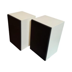 May include: Two white speakers with black front panels and a black trim. The speakers are rectangular with a slightly angled design. The speakers are placed on a white background.