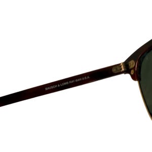 May include: Close-up of a pair of sunglasses with a dark brown frame and green lenses. The text "BAUSCH & LOMB RAY-BAN U.S.A." is printed on the arm of the sunglasses.