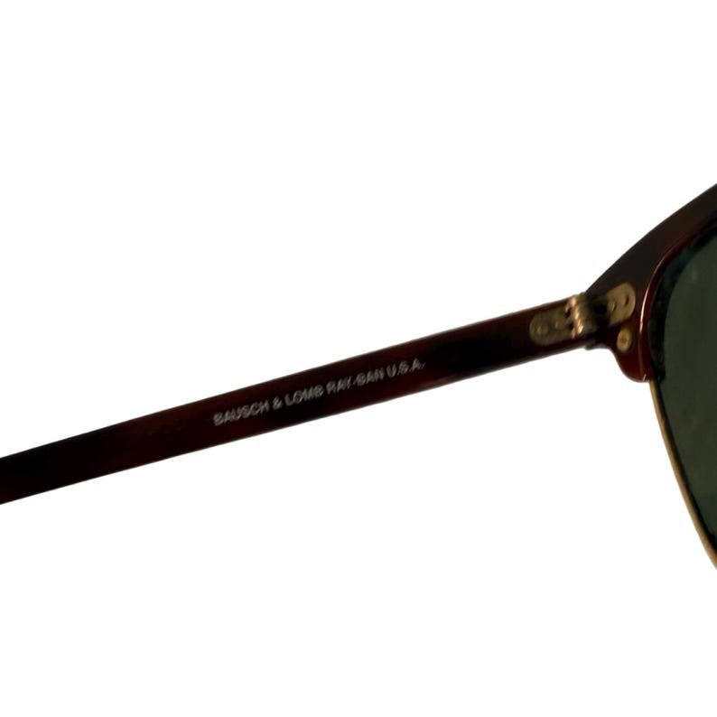 May include: Close-up of a pair of sunglasses with a dark brown frame and green lenses. The text "BAUSCH & LOMB RAY-BAN U.S.A." is printed on the arm of the sunglasses.