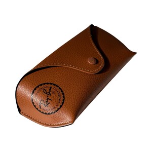 May include: A brown leather eyeglass case with a snap closure. The case has a rounded shape and features the Ray-Ban logo in black on the front. The case is designed to protect sunglasses.