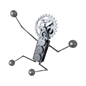 Op de afbeelding: Een grillige metalen sculptuur van een figuur geconstrueerd uit tandwielen, staven en bollen. Het hoofd is een groot tandwiel met wijzers, het lichaam is een rechthoekig frame met kleinere tandwielen, en de ledematen zijn staven met bollen aan de uiteinden.