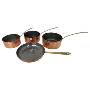 Op de afbeelding: Set van vier koperen potten met messing handvatten. De potten zijn van verschillende grootte en zijn in een halve cirkel gerangschikt. De grootste pot is een braadpan.