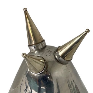 Pu&ograve; includere: Una latta per olio argentata con tre beccucci, ognuno con una punta appuntita. La latta ha una base arrotondata e una superficie liscia e riflettente.