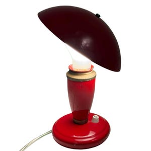 Op de afbeelding: Een vintage rode bureaulamp met een bordeauxrode kap. De lamp heeft een ronde voet, een cilindrisch lichaam en een gebogen kap. De gloeilamp is zichtbaar. De lamp heeft een schakelaar op de voet en een netsnoer.