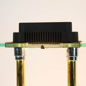 May include: A black electronic component with a grid-like pattern, supported by two gold and silver metal poles. The component sits on a clear glass surface, creating a modern, minimalist aesthetic.