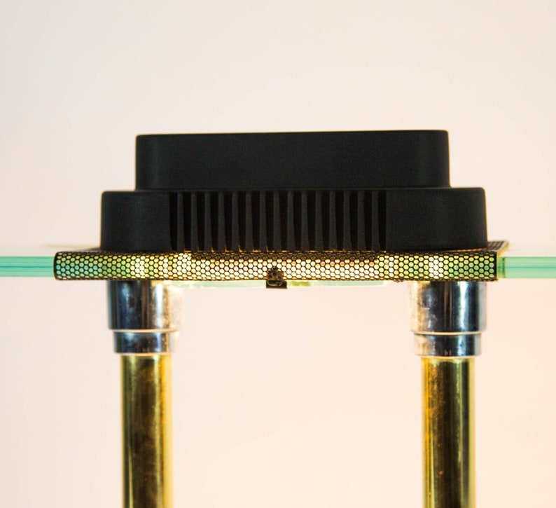 May include: A black electronic component with a grid-like pattern, supported by two gold and silver metal poles. The component sits on a clear glass surface, creating a modern, minimalist aesthetic.