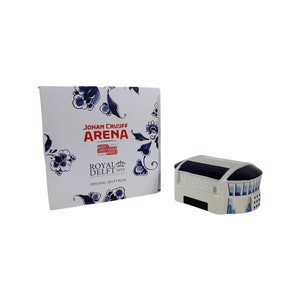 May include: A white ceramic model of the Johan Cruijff Arena, a famous football stadium in Amsterdam, Netherlands. The model is decorated with blue and white Delftware patterns, a traditional Dutch style of pottery. The box features the stadium's name and the Royal Delft logo.