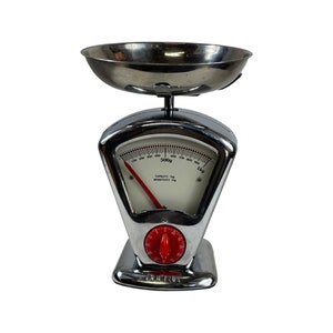 Puede incluir: Una báscula de cocina plateada con un dial rojo y un cuenco cromado. La báscula tiene una cara blanca con marcas negras y el texto "CAPACITY 1kg SENSITIVITY 10g" y "500g 1kg".