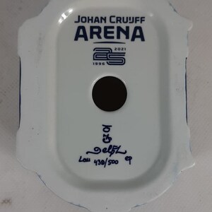 May include: White ceramic figurine of the Johan Cruijff Arena, a football stadium in Amsterdam, Netherlands. The figurine is decorated with blue details and the text "Johan Cruijff Arena" and "1996 2021". The figurine is numbered "439/500" and signed "Delft".