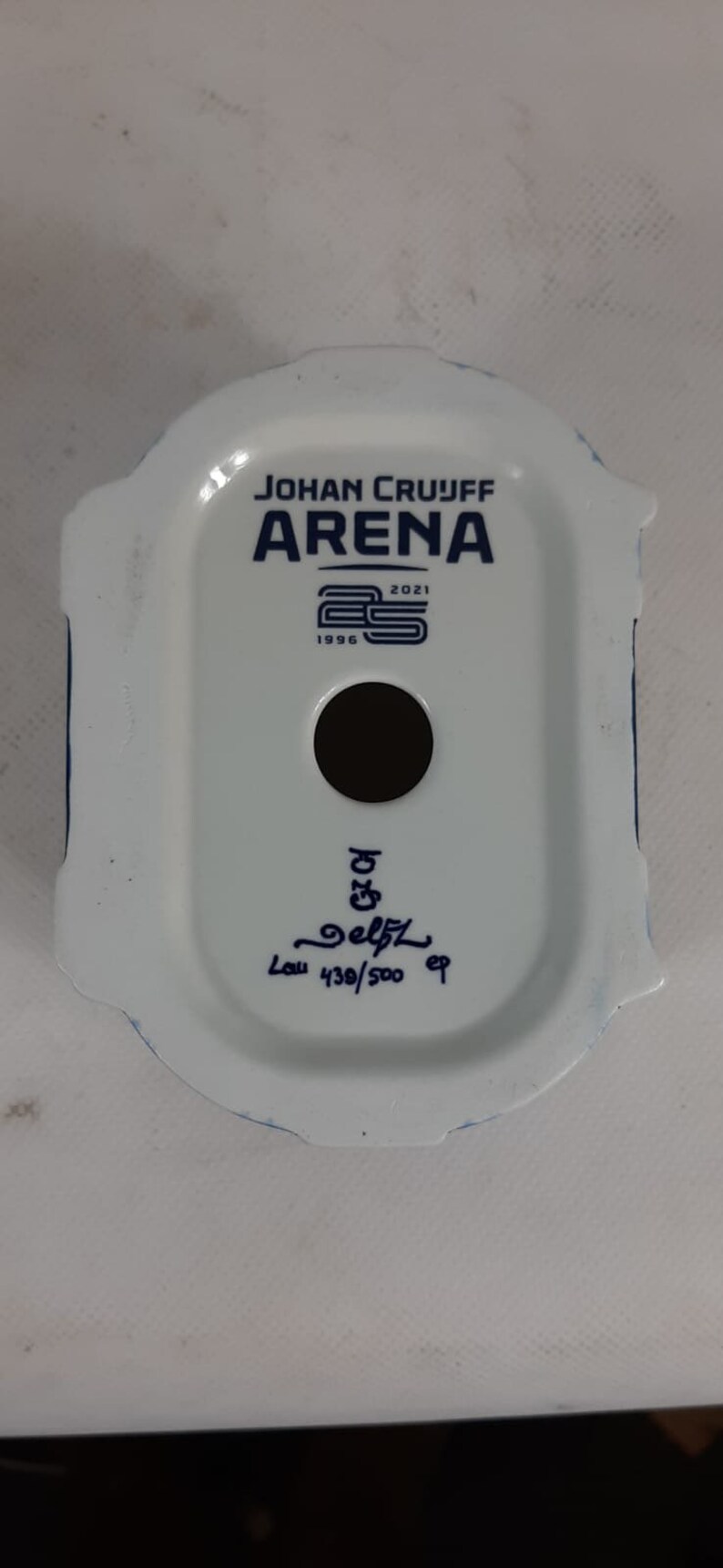 May include: White ceramic figurine of the Johan Cruijff Arena, a football stadium in Amsterdam, Netherlands. The figurine is decorated with blue details and the text "Johan Cruijff Arena" and "1996 2021". The figurine is numbered "439/500" and signed "Delft".