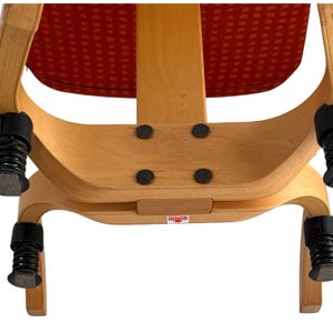 Puede incluir: Primer plano de una estructura de silla de madera con un asiento estampado en rojo. La silla tiene un dise&ntilde;o curvo con cuatro patas negras con resorte. El marco es de color marr&oacute;n claro y el asiento tiene un patr&oacute;n cuadrado repetitivo.