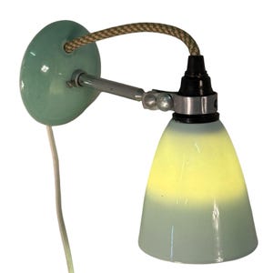 May include: A mint green wall sconce with a conical shade. The sconce features a circular base, a silver adjustable arm, and a textured brown and beige cord. The shade transitions from a pale green to a bright yellow.
