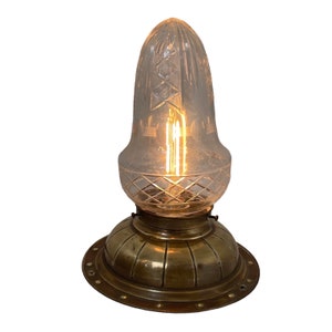 Puede incluir: Una lámpara de techo de latón y cristal con una pantalla de vidrio transparente facetado y una base redonda de latón. La luz está encendida y la bombilla es visible.