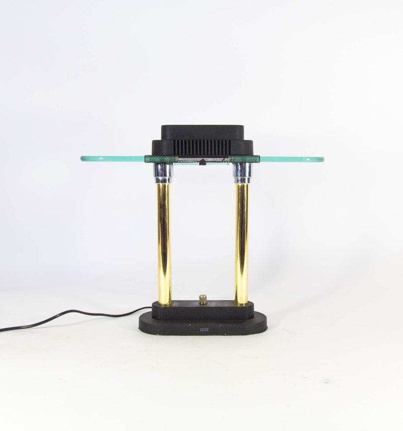May include: A modern desk lamp with a black rectangular top and base. The lamp features two gold-colored vertical supports and a clear glass shelf. The lamp is connected to a black power cord.