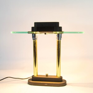 May include: A vintage desk lamp with a black rectangular top and a green glass shelf. The lamp features two gold-colored cylindrical supports and a brown base. The lamp is connected to a power cord.