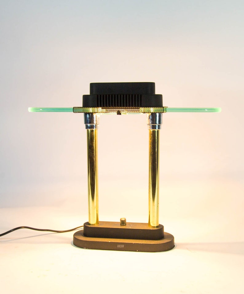 May include: A vintage desk lamp with a black rectangular top and a green glass shelf. The lamp features two gold-colored cylindrical supports and a brown base. The lamp is connected to a power cord.