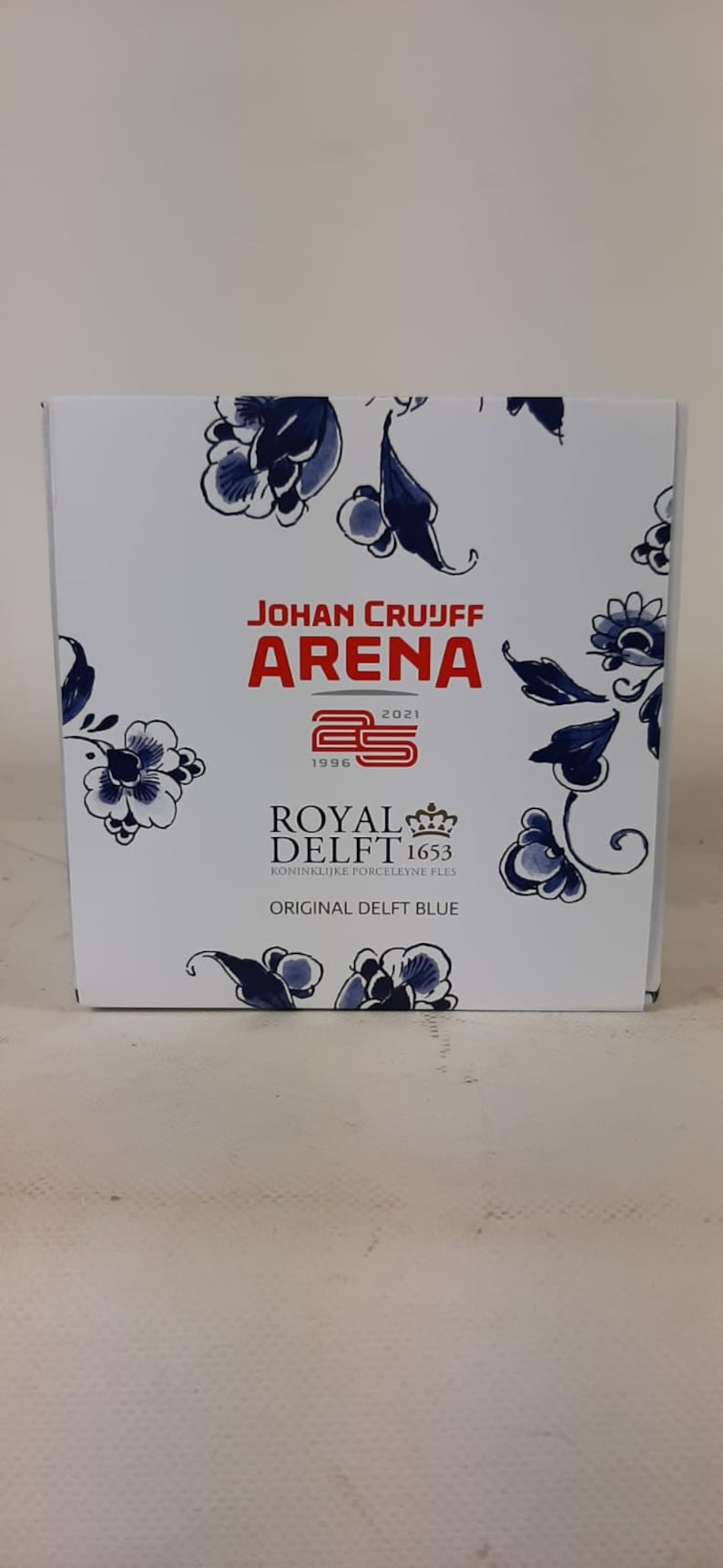 May include: A white box with a blue and white floral design. The box features the text "Johan Cruijff Arena 1996 2021" and a red logo with the number 25. The box also features the text "Royal Delft 1653 Koninklijke Porceleine Fles Original Delft Blue".