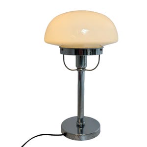 Può includere: Una lampada da tavolo cromata con un paralume in vetro bianco sporco a forma di fungo. La lampada ha una base e uno stelo cilindrici, con un supporto curvo che sostiene il paralume. Un cavo di alimentazione nero esce dalla base.