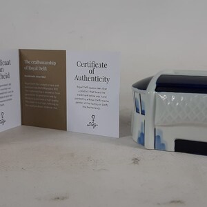 May include: A white and blue ceramic miniature of a stadium with a blue roof. The stadium has a white banner with a blue design. The miniature is accompanied by three certificates of authenticity from Royal Delft.