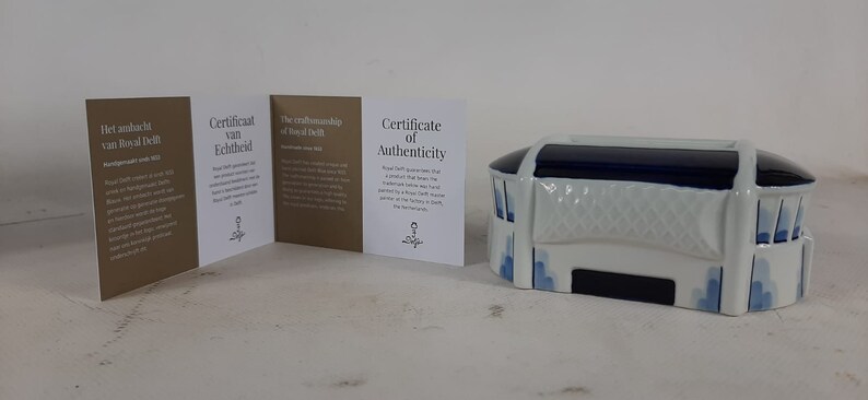May include: A white and blue ceramic miniature of a stadium with a blue roof. The stadium has a white banner with a blue design. The miniature is accompanied by three certificates of authenticity from Royal Delft.