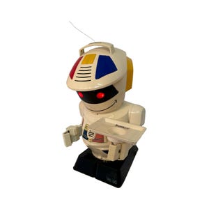 Op de afbeelding: Een vintage witte robotspeelgoed met een dienblad, rode ogen en een lachend gezicht. De kop van de robot heeft een handvat en is versierd met rode, gele en blauwe accenten. De robot staat op een zwarte basis.