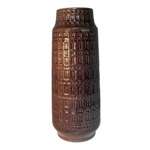 May include: A tall, brown ceramic vase with a textured, geometric pattern. The vase has a cylindrical shape with a slightly flared base and a narrower neck. The surface is covered in a repeating pattern of rectangles and circles. A small label is visible near the top.