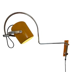 Puede incluir: Lámpara de pared de color amarillo mostaza con una pantalla cilíndrica y un brazo curvo de color plateado. La lámpara tiene una base de madera rectangular y un cable de alimentación negro. La lámpara es ajustable y direccional.