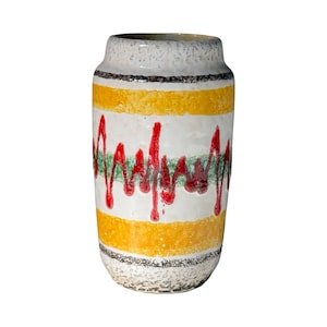 Puede incluir: Un jarrón cilíndrico de cerámica con una superficie blanca texturizada. El jarrón presenta bandas horizontales amarillas y blancas, con un diseño ondulado rojo y verde. Los bordes superior e inferior tienen detalles en negro y gris.