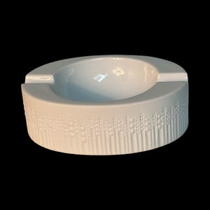 Op de afbeelding: Een witte keramische asbak met een gebogen ontwerp. De asbak heeft een verzonken gedeelte voor het vasthouden van sigaretten en een decoratief patroon van cirkels en lijnen rond de basis. De asbak staat op een zwarte achtergrond.