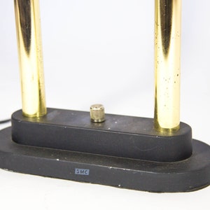May include: A black desk lamp with two vertical gold-colored metal tubes. The lamp has a black base with a small, cylindrical knob and a power cord. The base has the letters "SMC" printed on it.