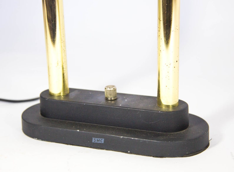 May include: A black desk lamp with two vertical gold-colored metal tubes. The lamp has a black base with a small, cylindrical knob and a power cord. The base has the letters "SMC" printed on it.