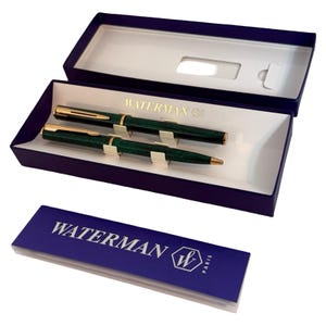 Pode incluir: Um conjunto de caneta e lapiseira Waterman numa caixa de apresentação azul. As canetas são verde escuro com detalhes dourados. A caixa tem o logótipo e o texto Waterman na tampa.