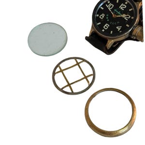 Könnte beinhalten: Eine Vintage-Armbanduhr mit schwarzem Zifferblatt, weißen Ziffern und einem schwarzen Lederarmband. Die Uhr hat ein goldfarbenes Gehäuse und wird von Uhrenreparaturteilen begleitet, darunter eine klare Glaslinse, ein Metallgitter und eine goldfarbene Lünette.