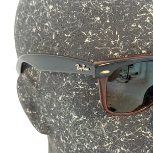 Może przedstawiać: Czarne i brązowe okulary przeciwsłoneczne z logo Ray-Ban na zauszniku. Okulary mają klasyczny design z czarną oprawką i brązowymi zausznikami. Soczewki są odblaskowe, na tle teksturowanym.