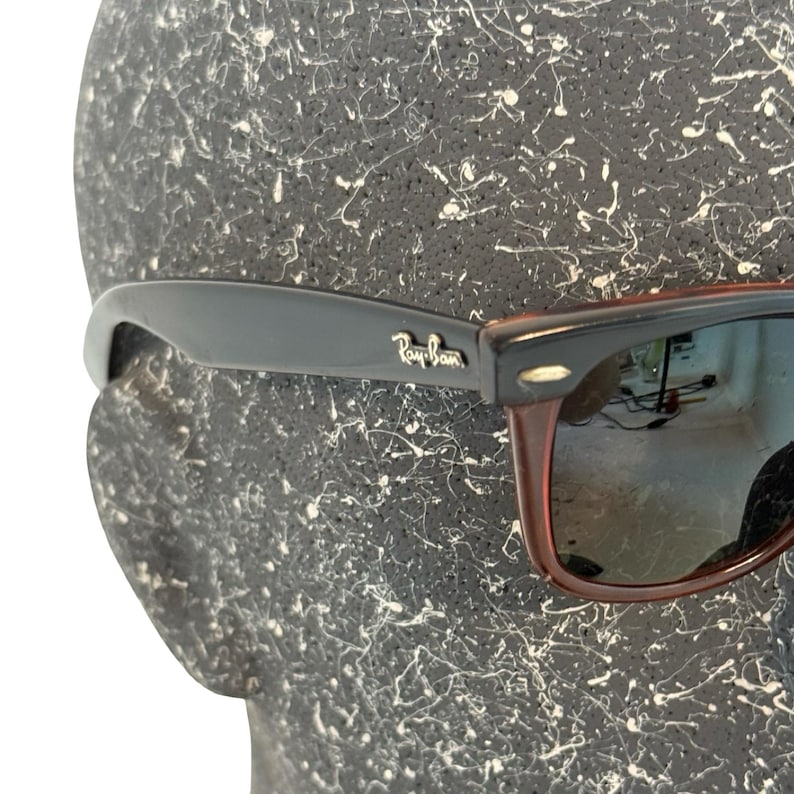 Może przedstawiać: Czarne i brązowe okulary przeciwsłoneczne z logo Ray-Ban na zauszniku. Okulary mają klasyczny design z czarną oprawką i brązowymi zausznikami. Soczewki są odblaskowe, na tle teksturowanym.