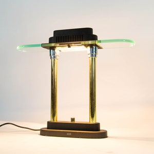 May include: A vintage desk lamp with a black rectangular top and a clear, oval-shaped glass shade. The lamp features two gold-colored cylindrical supports and a black base. The lamp is connected to a black power cord.