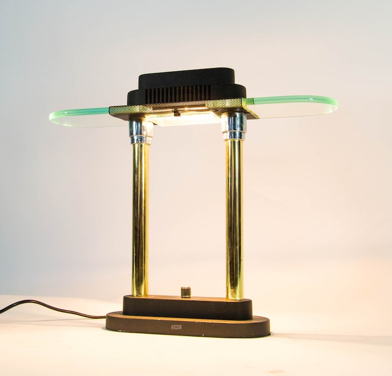 May include: A vintage desk lamp with a black rectangular top and a clear, oval-shaped glass shade. The lamp features two gold-colored cylindrical supports and a black base. The lamp is connected to a black power cord.