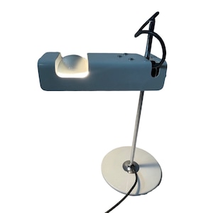 Puede incluir: Una lámpara de escritorio blanca con una pantalla redonda blanca y una base plateada. La lámpara tiene un cable negro y un brazo plateado que se extiende desde la base.