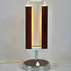 Könnte beinhalten: Eine moderne Tischlampe aus der Mitte des Jahrhunderts mit einem weißen Schirm und einem Sockel aus Holzmaserung. Die Lampe hat eine Chromoberfläche und einen runden Sockel.