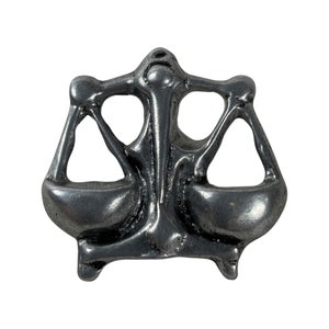 Op de afbeelding: Een zilverkleurige metalen hanger in de vorm van een weegschaal van de rechtvaardigheid. De weegschaal is in evenwicht en heeft een gedetailleerd ontwerp.