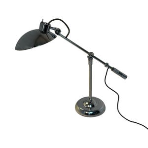 May include: A chrome-finished desk lamp with a curved shade and adjustable arm. The lamp has a round base and a black power cord. The shade is a dark, reflective color. The lamp is designed for task lighting.