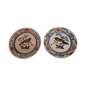Op de afbeelding: Twee keramische borden met een vogel- en bloemmotief. De borden hebben een crèmekleurig midden met een gedetailleerde illustratie van een vogel die op een tak zit. De randen zijn versierd met een blauwe rand en bloemmotieven in rood en oranje.