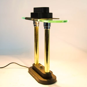 May include: A modern desk lamp with a black rectangular base and two gold-colored vertical supports. The lamp features a green glass shelf and a black, tiered top. The lamp is plugged into a wall outlet.