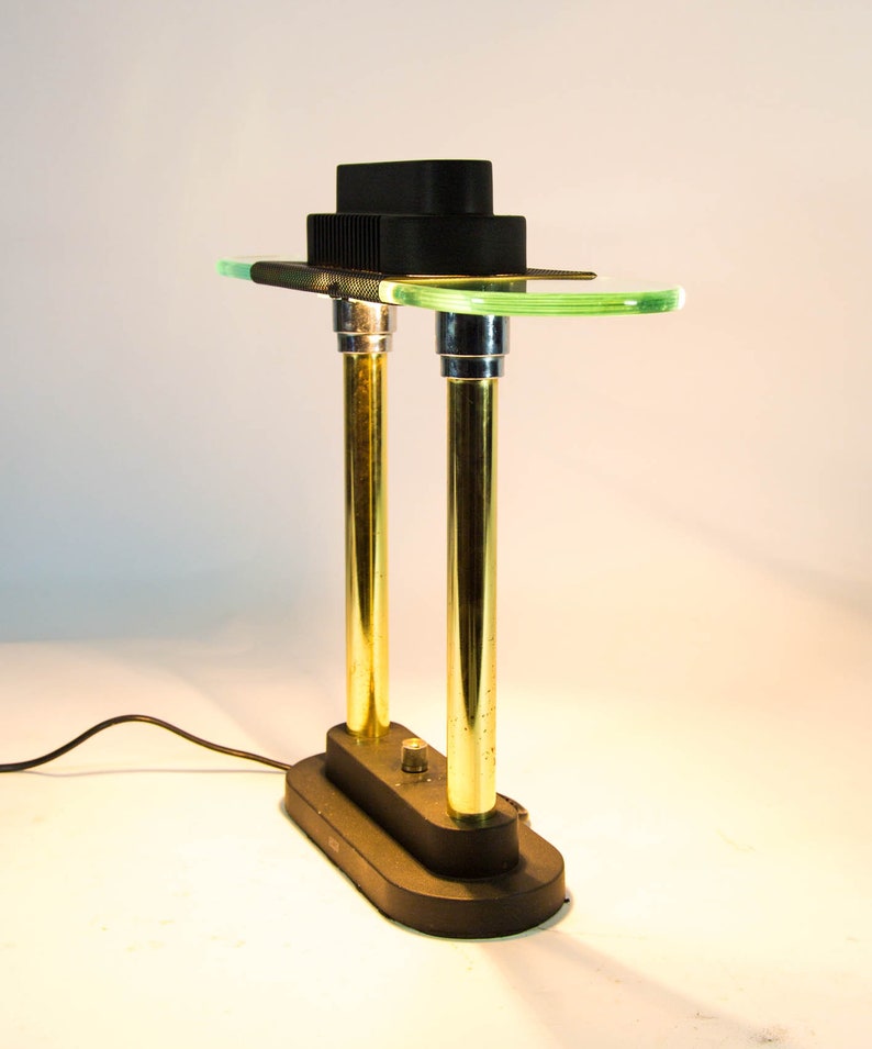May include: A modern desk lamp with a black rectangular base and two gold-colored vertical supports. The lamp features a green glass shelf and a black, tiered top. The lamp is plugged into a wall outlet.