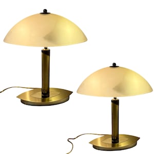 Op de afbeelding: Twee bijpassende tafellampen met een messing afwerking. Elke lamp heeft een halfronde, crèmekleurige kap en een cilindrische steel. De voet heeft een ovale vorm. De lampen zijn via een snoer aangesloten op een stroombron.