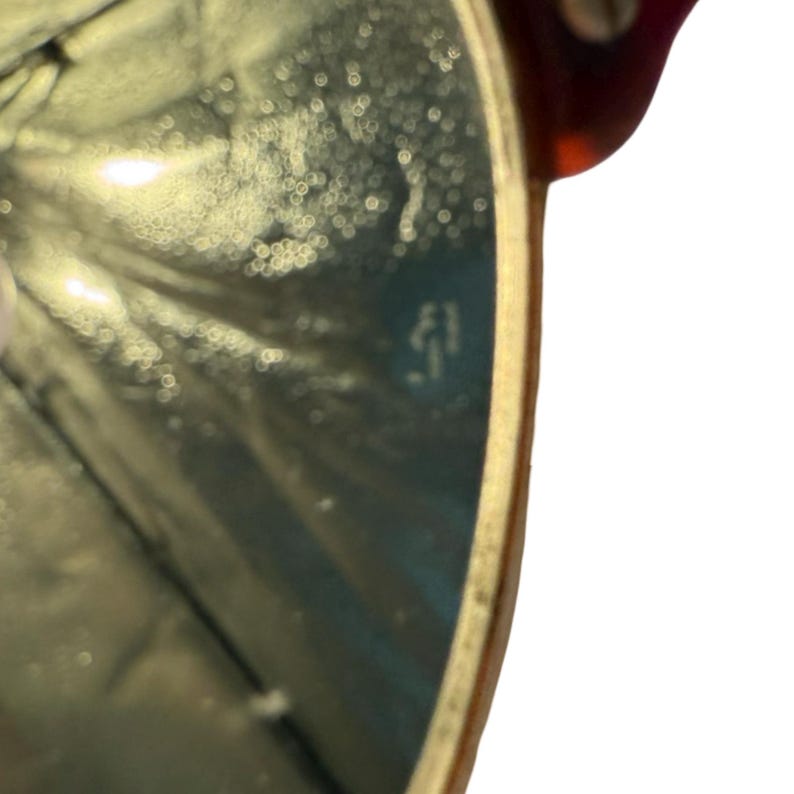 May include: Close-up of a gold-rimmed eyeglass lens with a reflective surface. The lens has a subtle, textured pattern and a small, etched detail near the center. The frame is a warm, reddish-brown color.