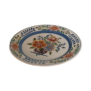 Op de afbeelding: Ovale keramische plaat met een bloemenontwerp. De plaat heeft een blauwe rand en een centrale opstelling van oranje, blauwe en witte bloemen. De rand is versierd met groene en blauwe bladerpatronen.