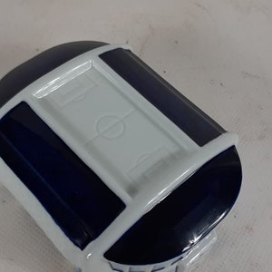 May include: A white ceramic trinket box shaped like a stadium with a blue trim and a white football pitch on the top.