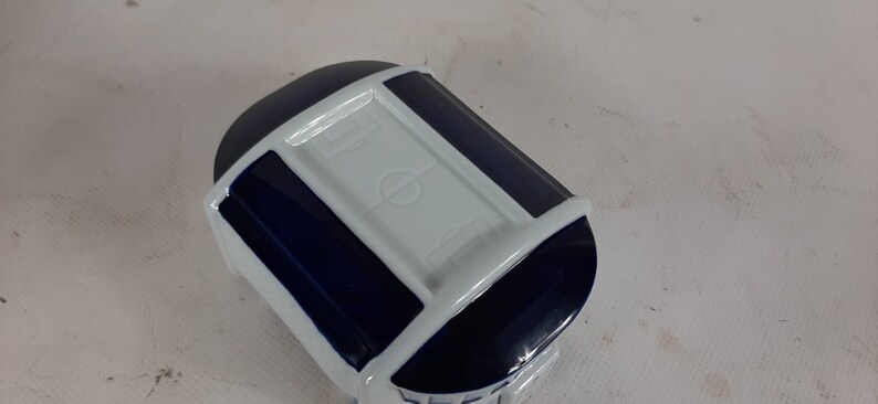 May include: A white ceramic trinket box shaped like a stadium with a blue trim and a white football pitch on the top.